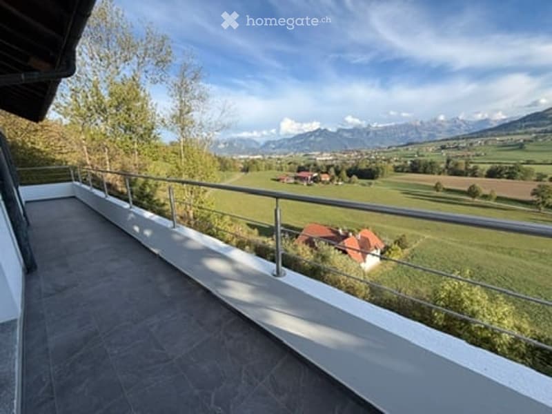 Balcony with metal railing overlooking a green field and distant mountains, with a house visible in the distance.