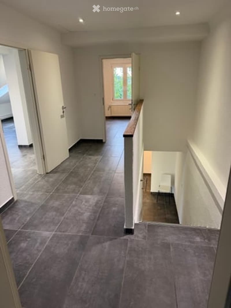 Spacious hallway with grey tiled flooring, white walls, and a wooden handrail leading to an upper level. Open doors reveal adjacent rooms.