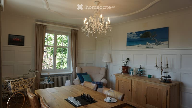 A well-furnished room with a wooden dining table, sofa, chandelier, and decorative items including a chessboard and a baby stroller.