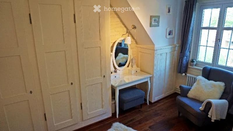 Cozy attic room with wooden floors, white built-in wardrobe, vanity table with mirror, armchair, throw pillow, and radiator.