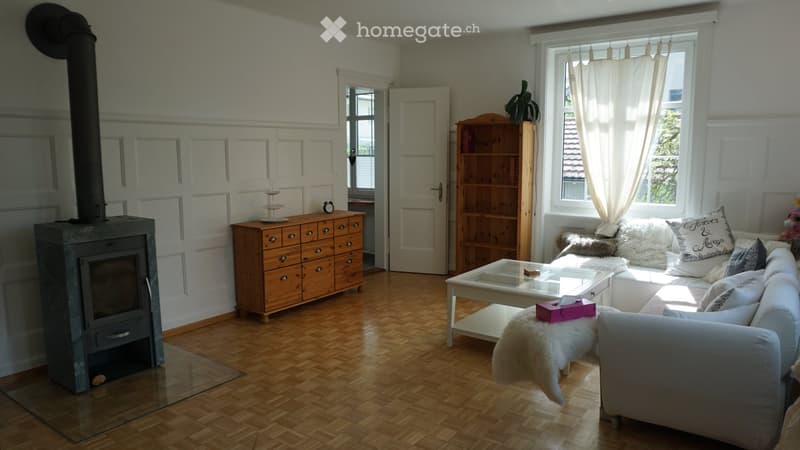 Cozy living room with wooden flooring, white walls, large windows with sheer curtains, a white sectional sofa with decorative pillows, a glass coffee table, a wooden cabinet, a bookshelf, and a stone fireplace.