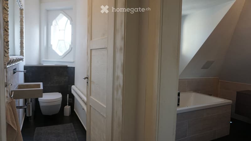 Modern bathroom with a sink, toilet, bathtub, and a mirror. The room features a sloped ceiling and a window with a decorative glass design.