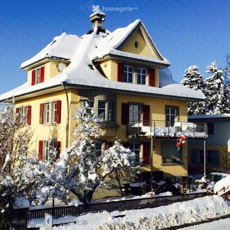 A two-story yellow house with red shutters and snow-covered roof and balcony. The house features multiple windows and a chimney, surrounded by snow-covered trees and a fence.