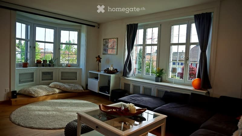 Bright living room with large windows, a dark grey sofa, a white coffee table, a round white rug, and a small table with a lamp and potted plants.