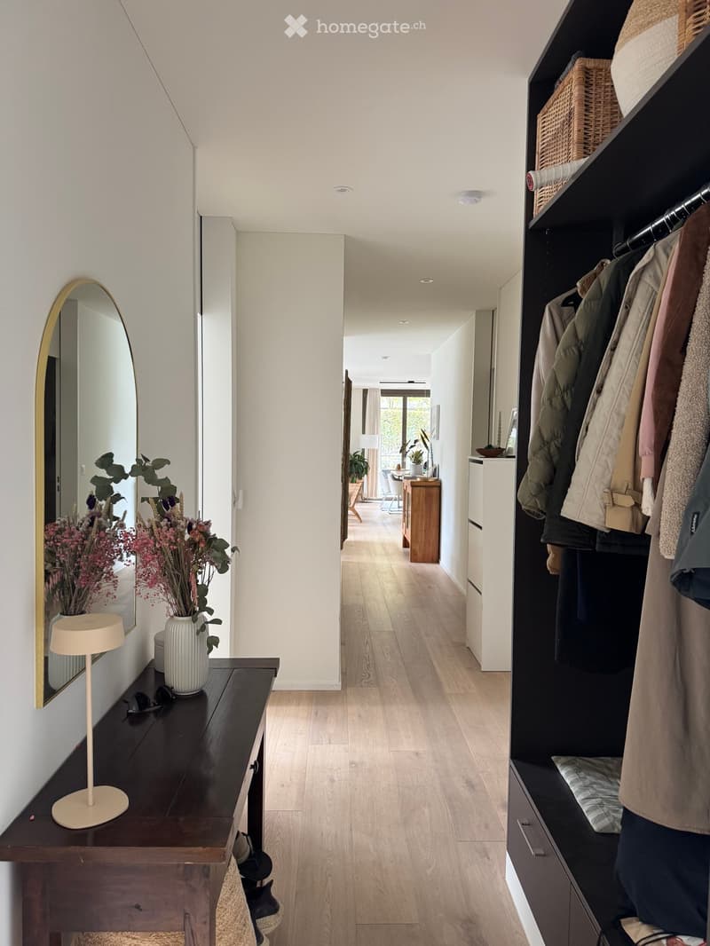 Modern hallway with light wooden flooring, built-in closet with hanging coats, small table with vase and lamp, and a mirror on the wall.