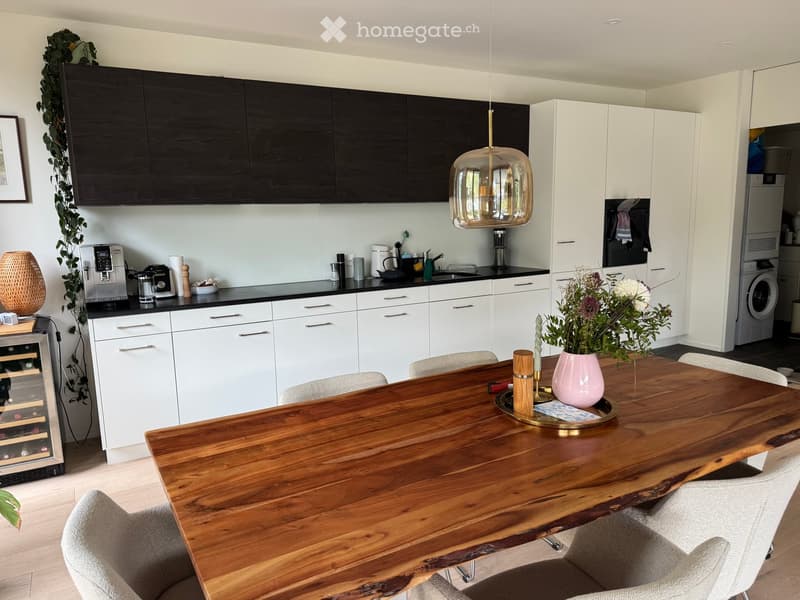 Modern kitchen with white and black cabinets, a large wooden dining table with chairs, a wine cooler, coffee maker, and a pendant light fixture.