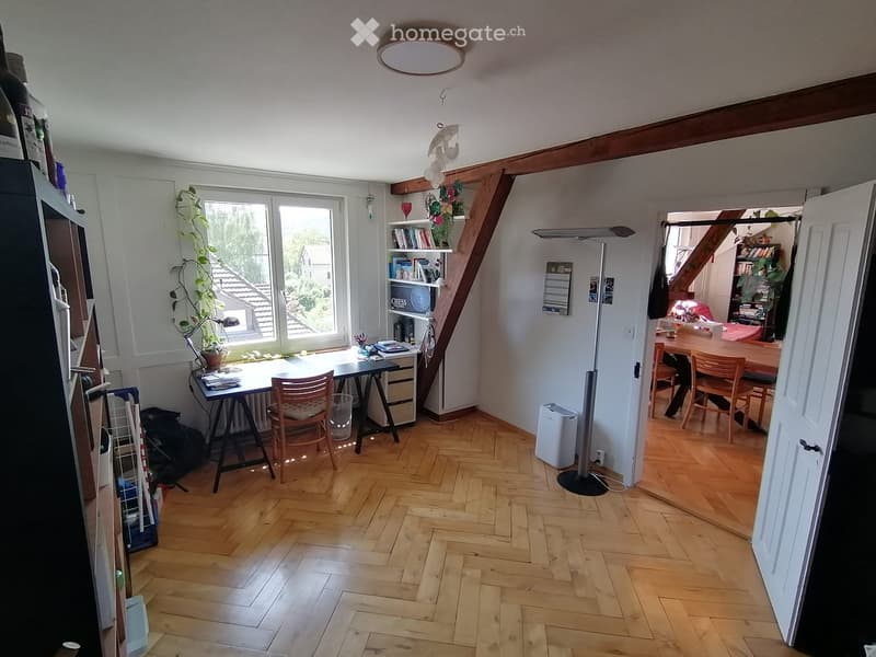 A well-lit room with wooden flooring, a large window, a desk with a chair, a bookshelf, a lamp, and a small dining area with a table and chairs.