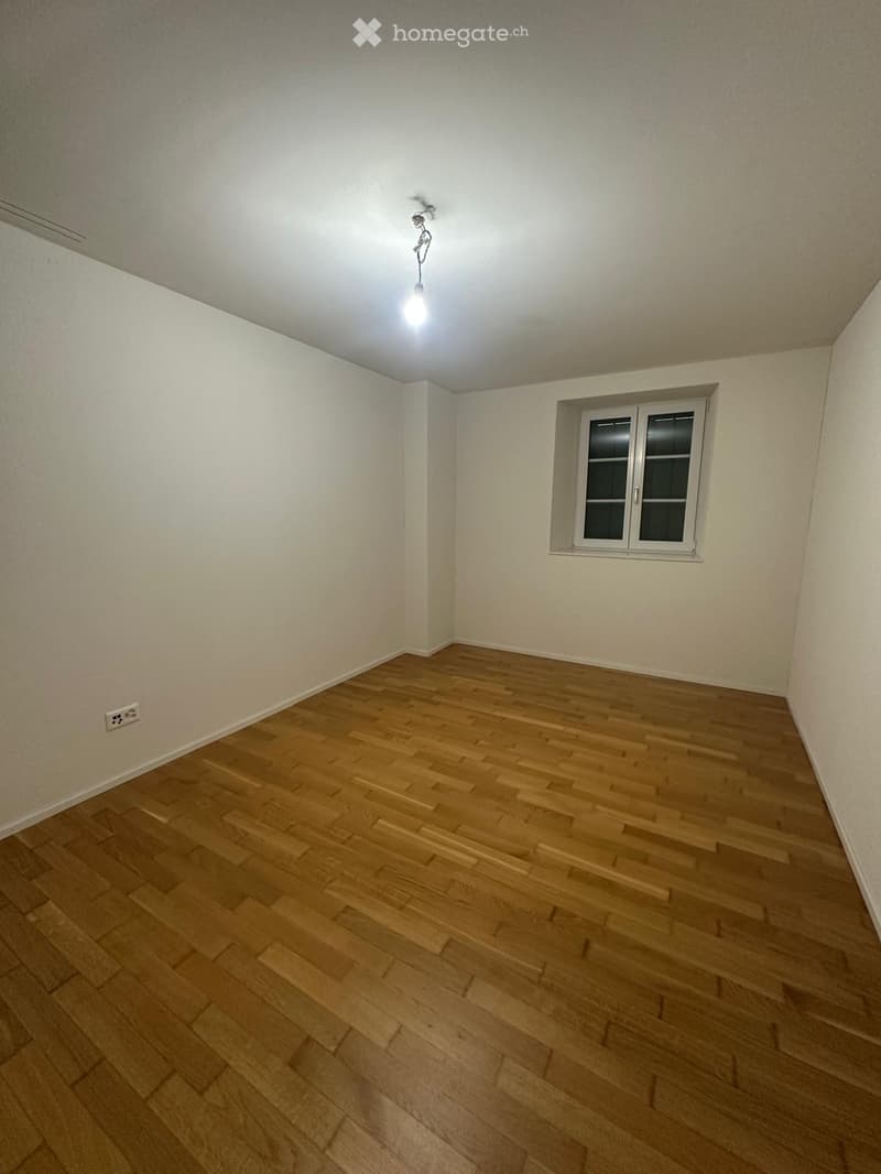 Empty room with wooden flooring, white walls, and a single window with white frame. A light fixture hangs from the ceiling.