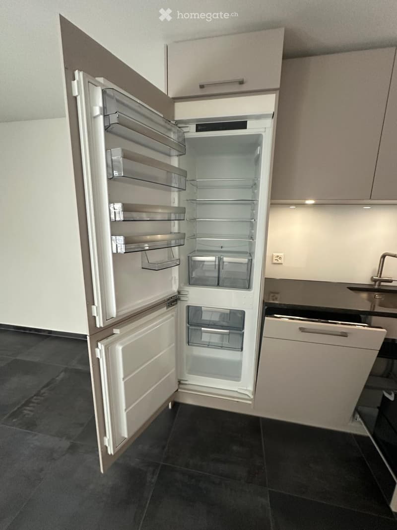 Modern kitchen with a stainless steel refrigerator, sink, and faucet, featuring light-colored cabinets and dark tiled flooring.
