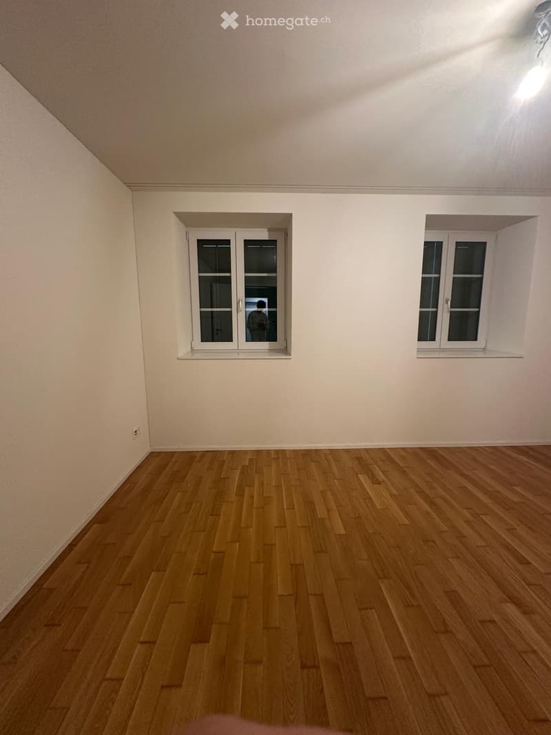 Empty room with wooden flooring and white walls, featuring two windows with white frames and a ceiling light fixture.