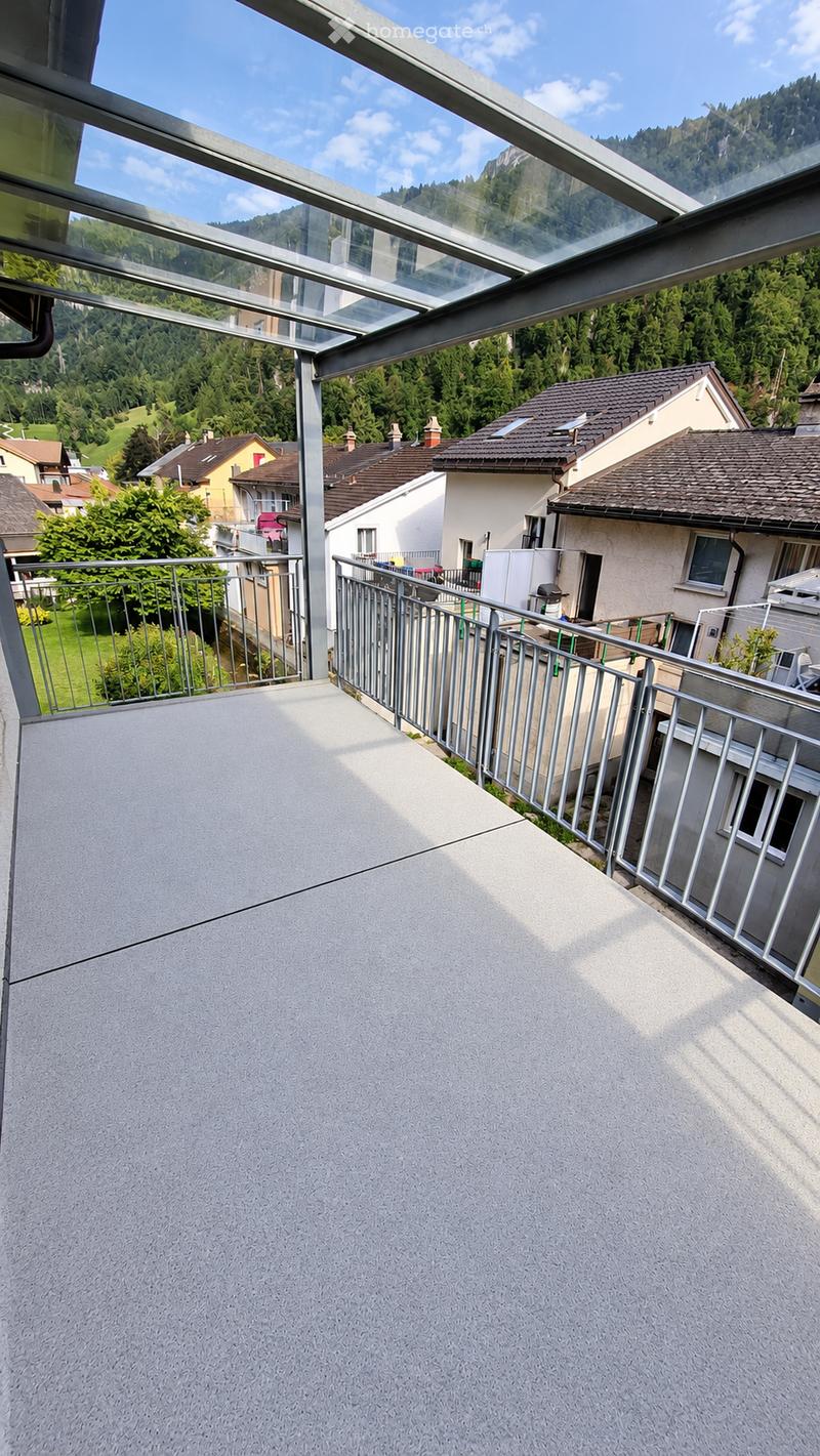 Balcony with a modern glass roof, metal railing, and a view of a green, hilly landscape with houses and trees.