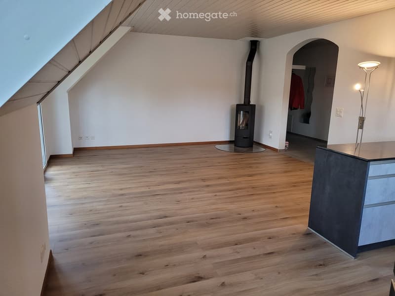 Empty attic room with wooden flooring, white walls, and a fireplace in the corner. The room features exposed wooden beams on the ceiling and a large skylight.