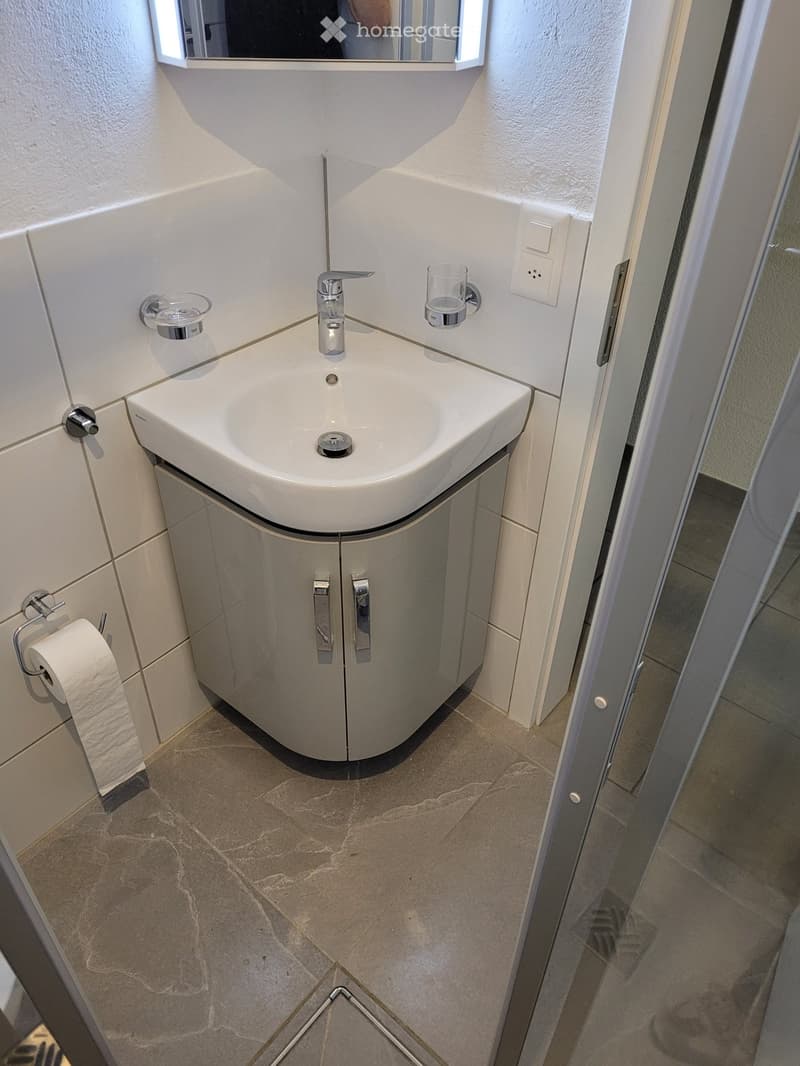 Modern bathroom with a white sink, chrome faucet, mirrored cabinet, and a small storage cabinet beneath the sink. The floor is tiled with beige stone tiles.