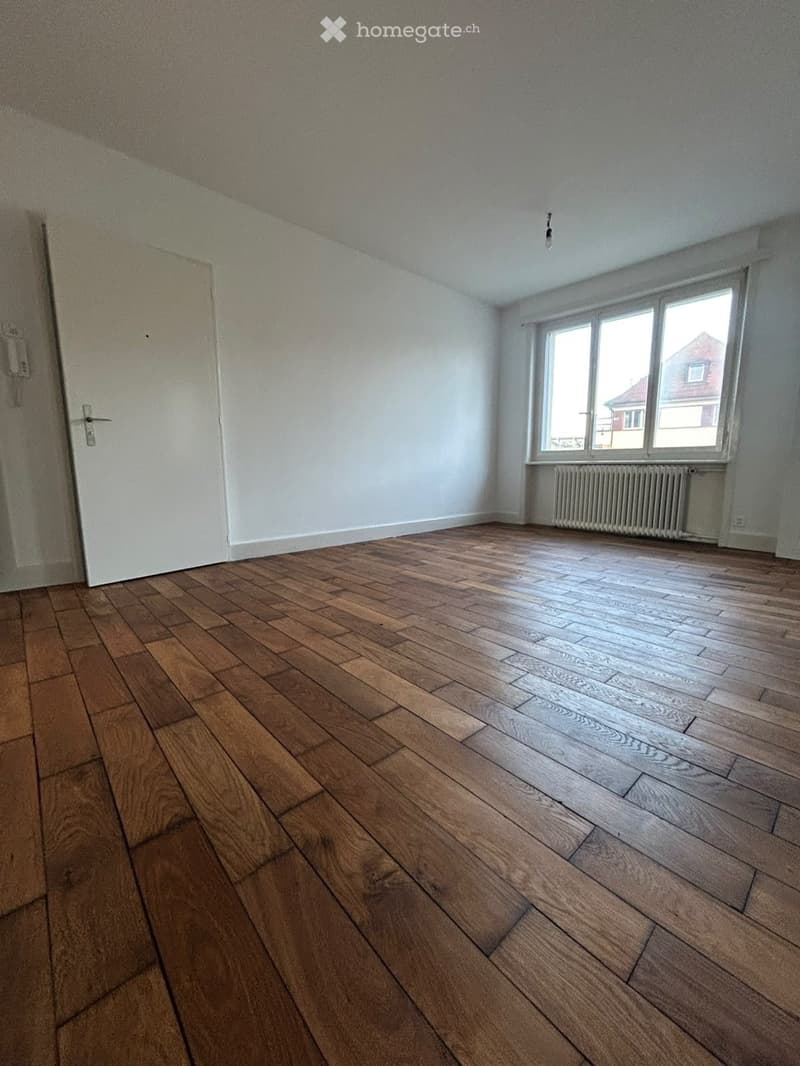 Empty room with wooden flooring, white walls, a window with white frame, and a radiator below it. A white door is visible on the left side.