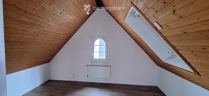Empty attic room with wooden ceiling, white walls, wooden floor, and a small window with a white frame.