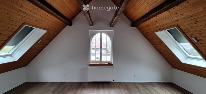Empty attic room with wooden ceiling beams, wooden flooring, and multiple windows including a central arched window and two skylights.
