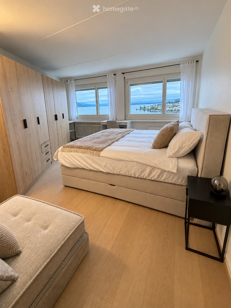 Bright bedroom with wooden flooring, large windows offering a scenic view, a neatly made bed with pillows and a blanket, a nightstand with a lamp, a chair, and a desk near the window.