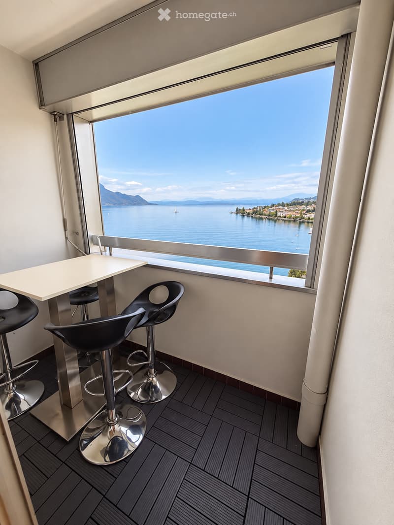 Modern balcony area with a view of a lake and mountains, featuring a white table and black stools.