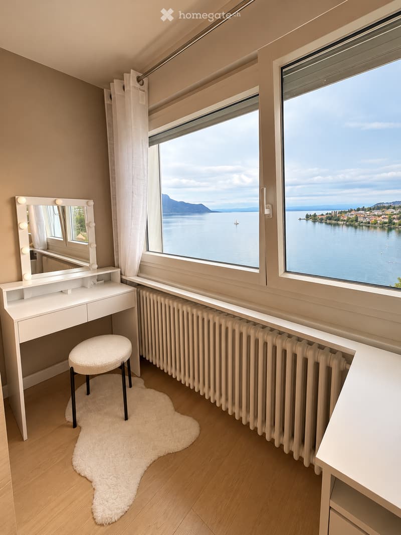 Well-lit room with large windows overlooking a lake, featuring a vanity with a mirror, a white stool, a radiator, and a fluffy white rug on the wooden floor.