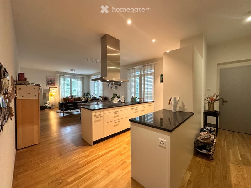 Open-plan kitchen and living area with light wood flooring, modern kitchen island, refrigerator, sofa, and framed pictures on the wall.