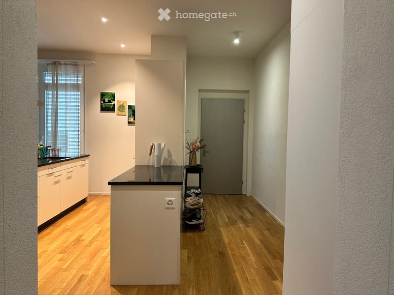 A modern hallway with wooden flooring, white walls, and a small kitchen area visible on the left. The hallway features a closed door at the end and a small table with shoes and a vase of flowers near the door.