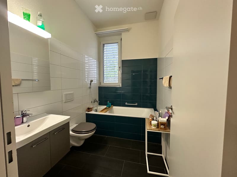 Modern bathroom with white tiles, dark blue accent wall, bathtub, toilet, sink, and mirror. Features a window with blinds and a small shelf with toiletries.
