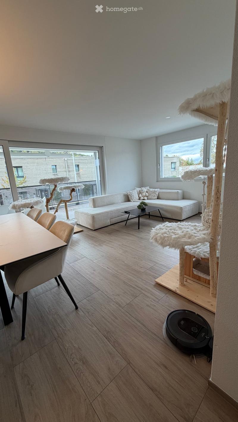Modern living room with light wooden flooring, white walls, large windows, and a cat tree. Features a white sectional sofa, coffee table, dining table with chairs, and a robotic vacuum cleaner.