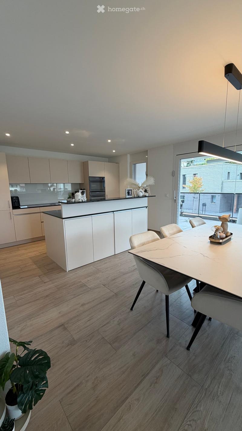 Modern kitchen and dining area with light-colored cabinets, a central island, and a dining table with chairs. The space features wooden flooring and large windows with a view to an outdoor area.