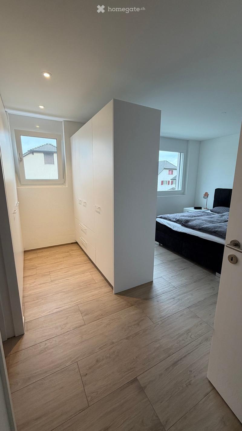 Modern bedroom with wooden flooring, large windows, a bed with dark headboard, nightstand, lamp, and a white wardrobe with drawers.