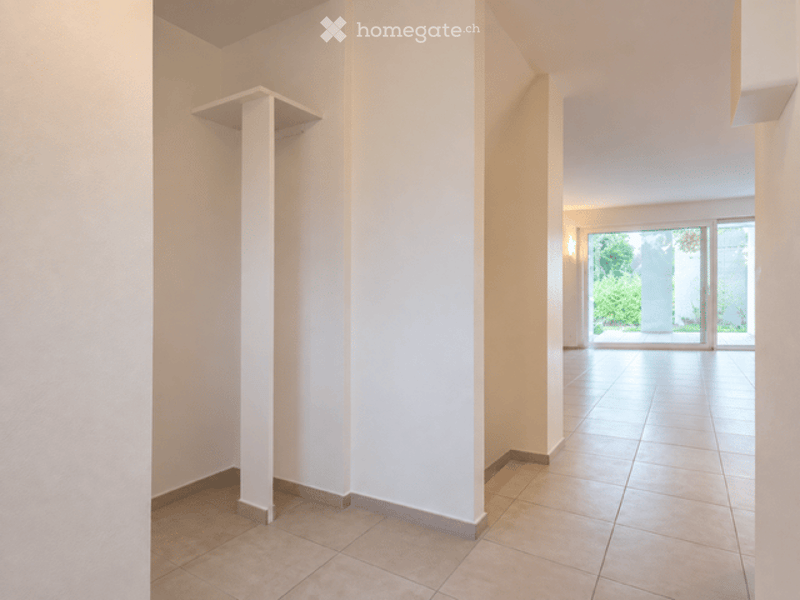 Empty hallway with white walls, tiled floor, and a built-in shelf on one side. The hallway leads to an open area with large glass doors and a view of greenery outside.