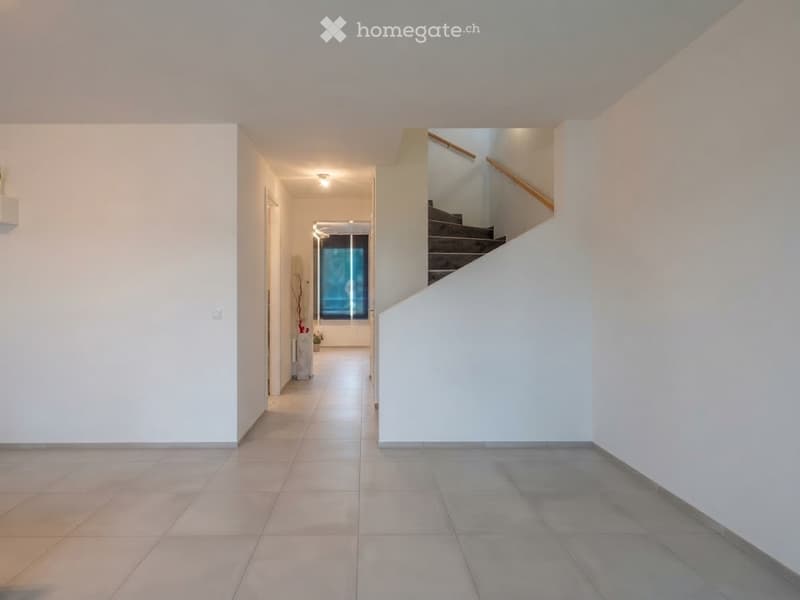 Bright hallway with tiled flooring, white walls, and a staircase leading upwards. The hallway features a small decorative plant and a window at the end, allowing natural light to enter.