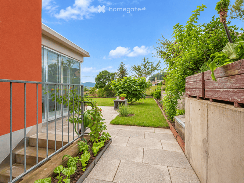 A well-maintained garden area with a paved pathway, green lawn, various plants, and a small seating area with a table and chairs.