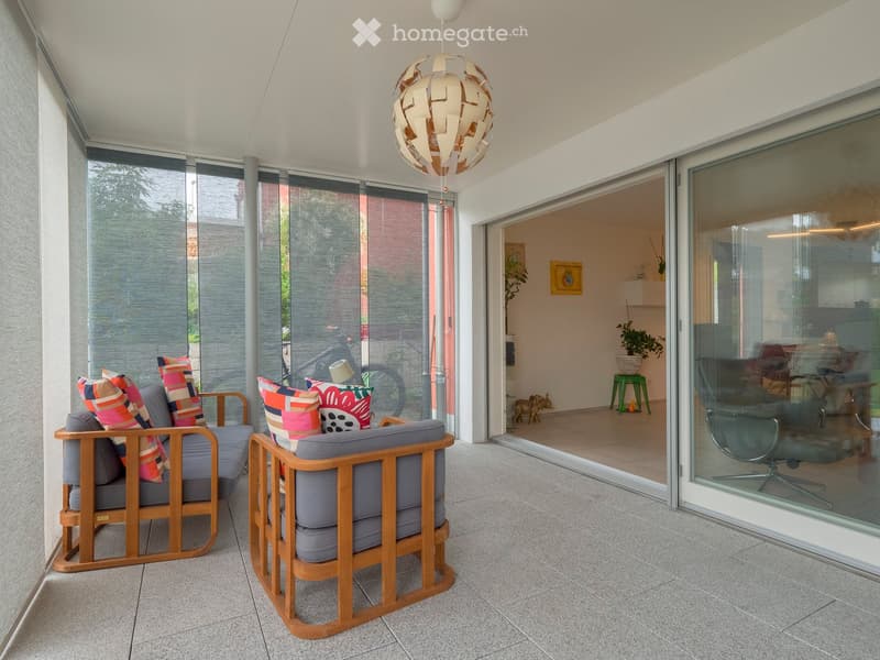 Modern balcony with large glass doors, two wooden armchairs with colorful cushions, a hanging lamp, and a view of the interior living space.