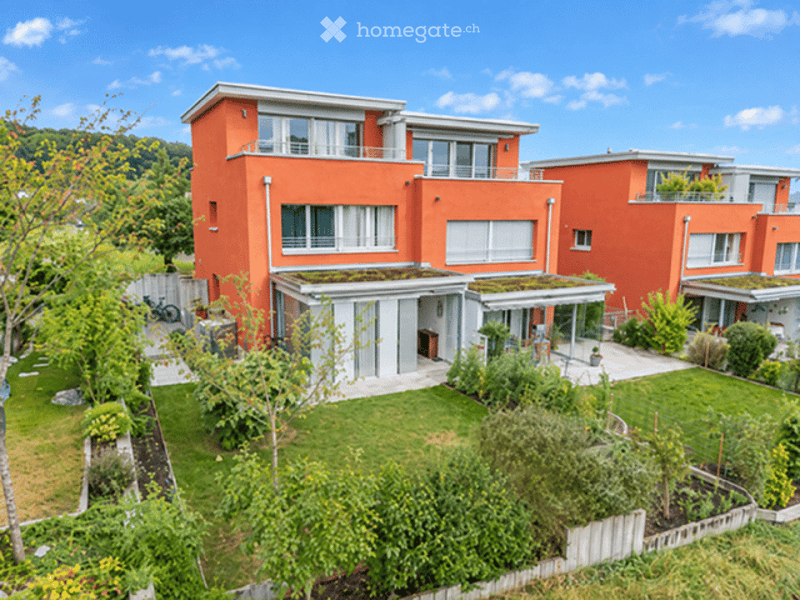 Modern two-story house with orange exterior, white trim, large windows, and a green roof. The house features a well-maintained garden with lawn, trees, and a patio area.