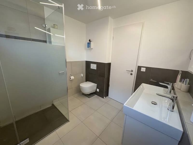 Modern bathroom with white tiles, a glass-enclosed shower, a toilet, a sink with a modern faucet, and a toothbrush holder with toothpaste on the counter.