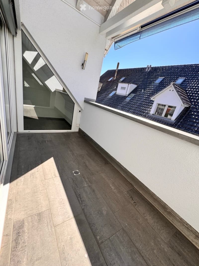 Balcony with wooden decking, white walls, large windows, and a view of the roof and neighboring buildings.
