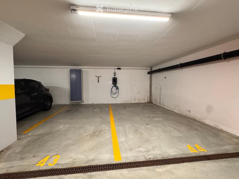 Underground parking space with a single car parked, marked with yellow lines and numbers, featuring a charging station and a storage cabinet.