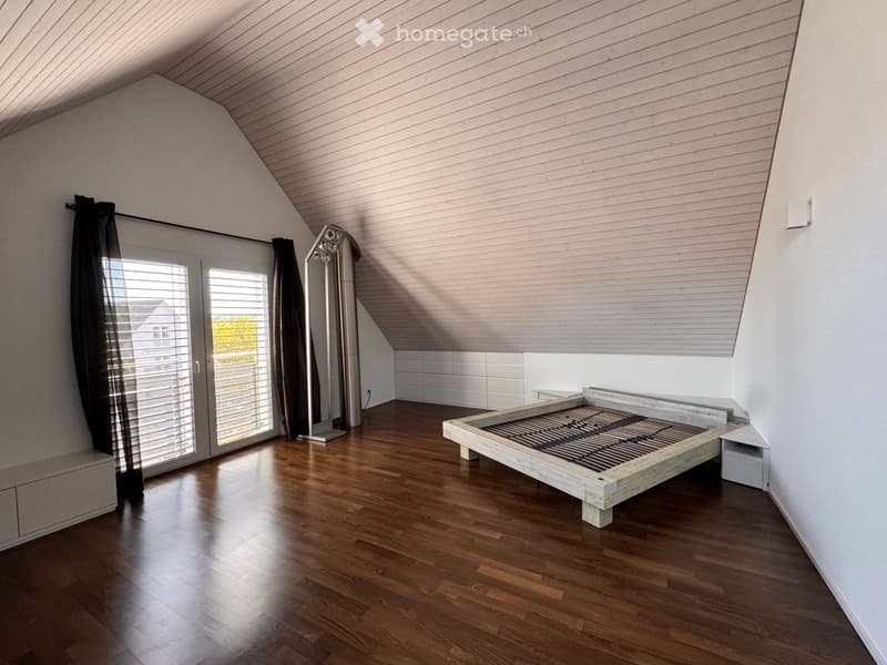 Bright, modern bedroom with wooden flooring, a simple wooden bed frame, a nightstand, a large mirror, and large windows with curtains.