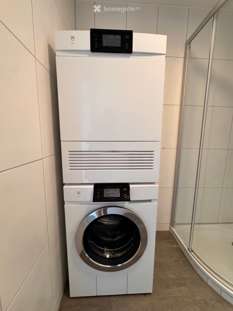 Modern bathroom with white tiled walls, a glass-enclosed shower, and stacked white washing machine and dishwasher units.