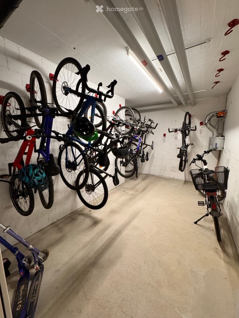 A well-lit indoor corridor with concrete floors and white brick walls, featuring multiple bicycles mounted on racks along the walls, with helmets and accessories visible.