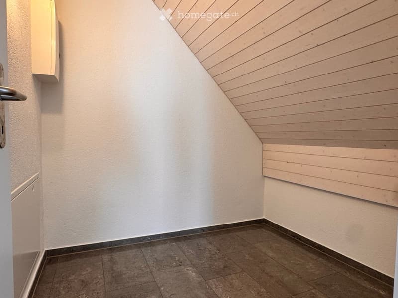 Empty hallway with white walls, wooden slanted ceiling, tiled floor, and a door with a silver handle on the left side.
