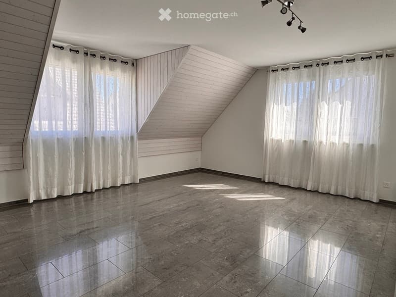 Empty attic room with slanted wooden ceiling, large windows covered by white curtains, and polished tile flooring.