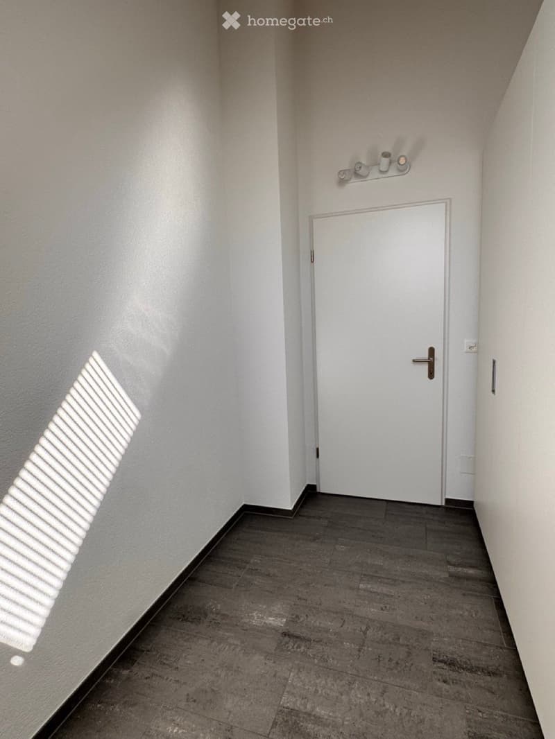 Empty hallway with a white door at the end, dark tiled floor, and a wall-mounted light fixture above the door.