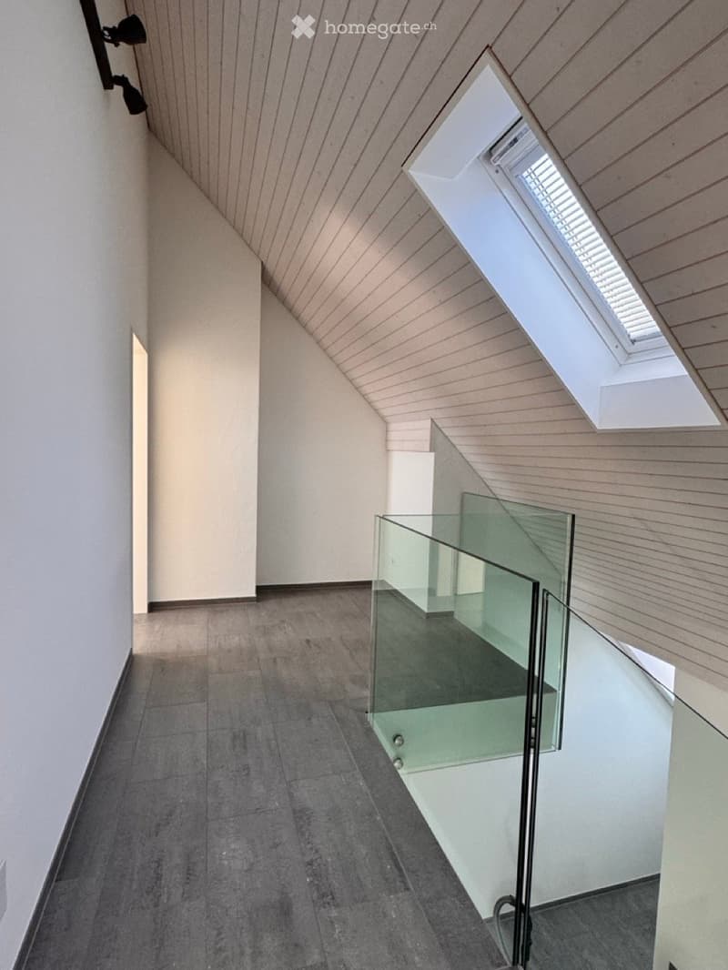 Modern hallway with wooden slatted ceiling, grey tiled floor, and glass railing leading to a bright skylight window.