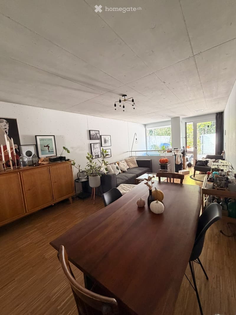 Modern living and dining area with wooden flooring, exposed concrete ceiling, large windows, and a view of the outdoor space. Features include a grey sectional sofa, wooden dining table with chairs, a wooden cabinet with decorative items, and several pott