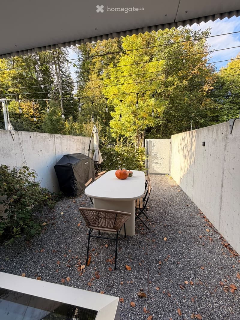 Compact outdoor patio with a white table, chairs, a covered grill, and string lights, surrounded by concrete walls and greenery.