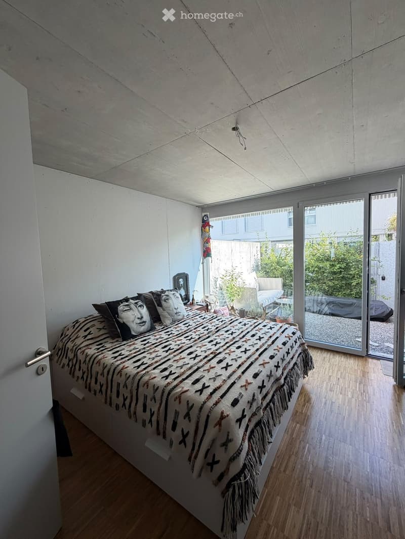 Modern bedroom with wooden flooring, large bed with patterned bedspread and pillows, nightstand, mirror, and a sliding glass door leading to a patio area.