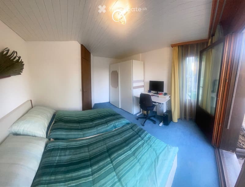 Cozy bedroom with two single beds, a desk with a computer, a chair, and a wardrobe. The room features blue carpeting and white walls.