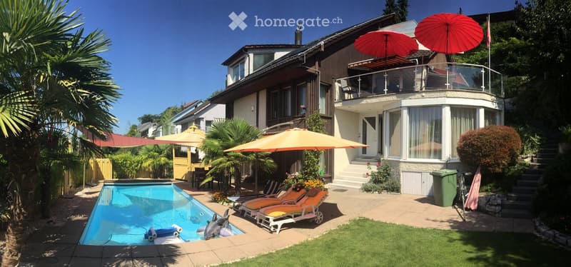A modern two-story house with a spacious backyard featuring a swimming pool, lounge chairs, umbrellas, and a well-maintained garden with palm trees and shrubs.