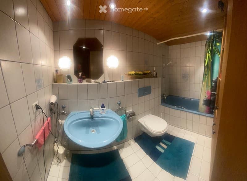 A small bathroom with a blue sink, white toilet, blue bathtub, and a large mirror above the sink. The walls are tiled, and there are two blue rugs on the floor.